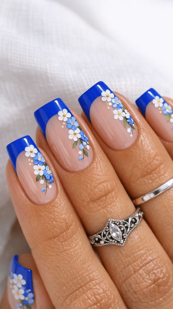 Square nails with bold blue French tips and cascading forget-me-not flowers in blue and white with green leaves on a nude base