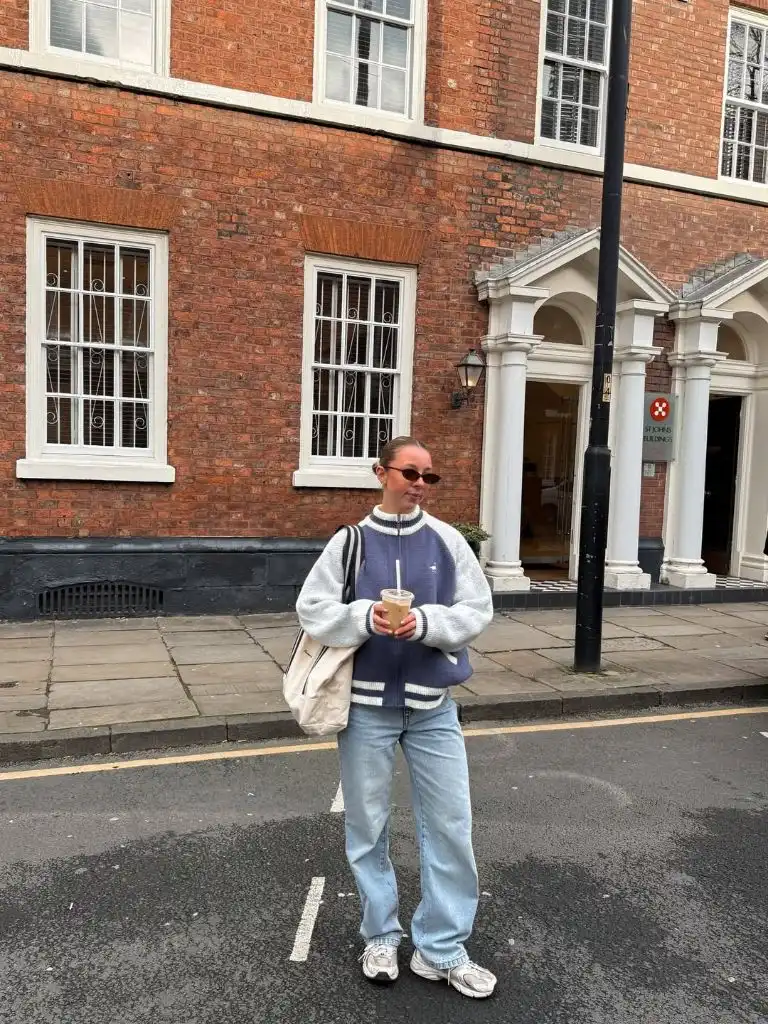 Navy and grey varsity-style zip jacket with light-wash straight-leg jeans, white sneakers, and canvas tote