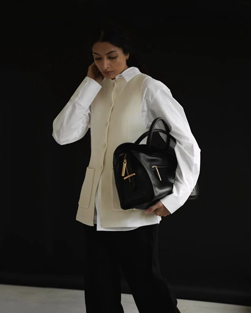 Beige knit button-front vest over white oversized shirt with black trousers and structured black leather bag