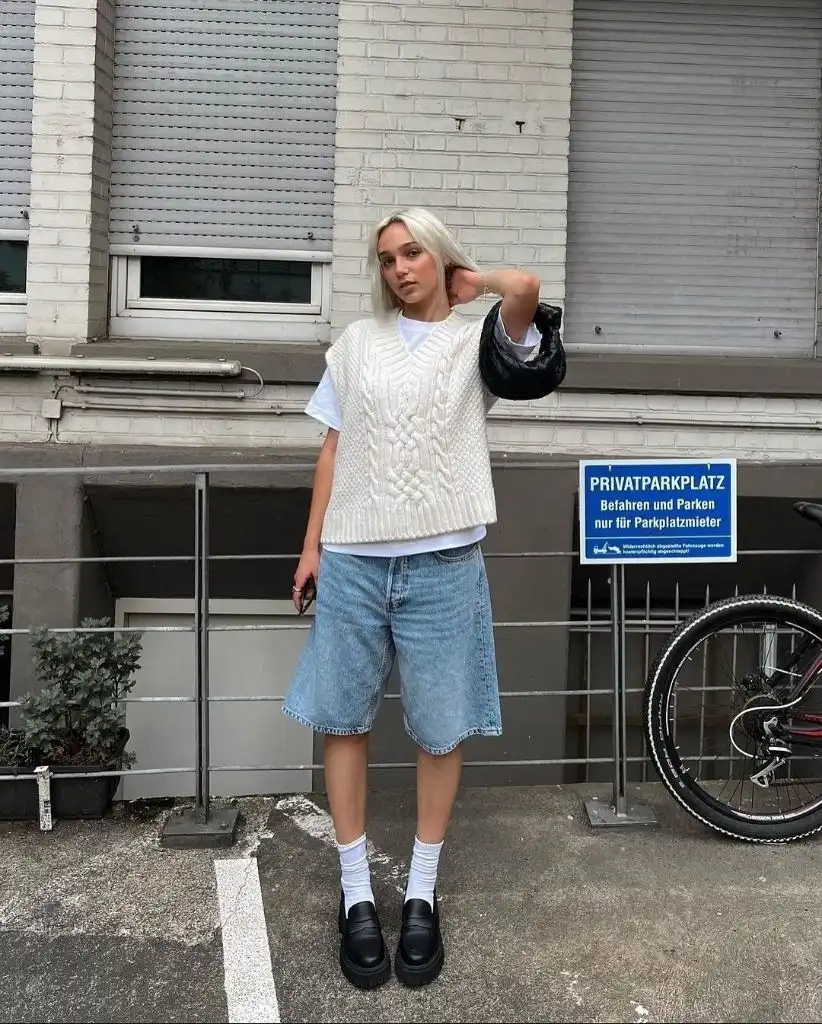 White oversized cable knit vest over white t-shirt with light wash denim bermuda shorts, white crew socks, and black chunky loafers