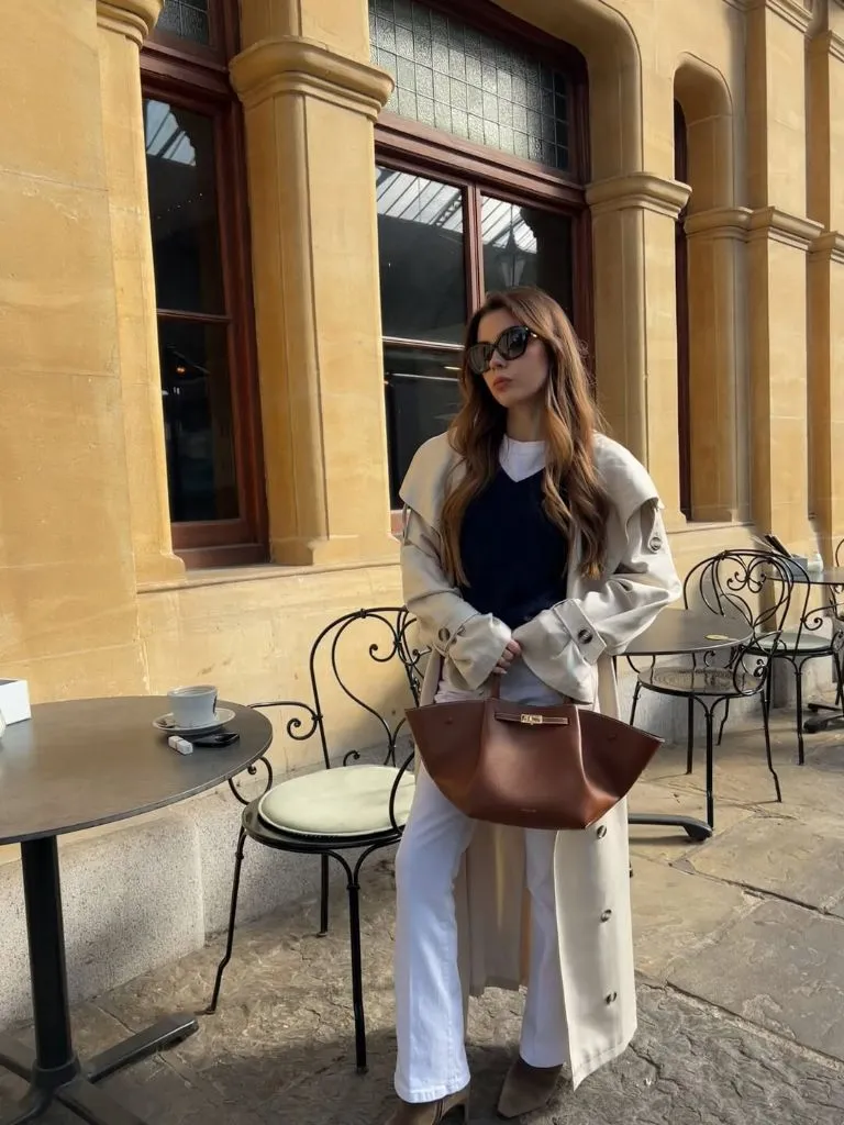 Cream trench coat layered over navy V-neck sweater, white tee, white wide-leg pants, and cognac leather tote at European café