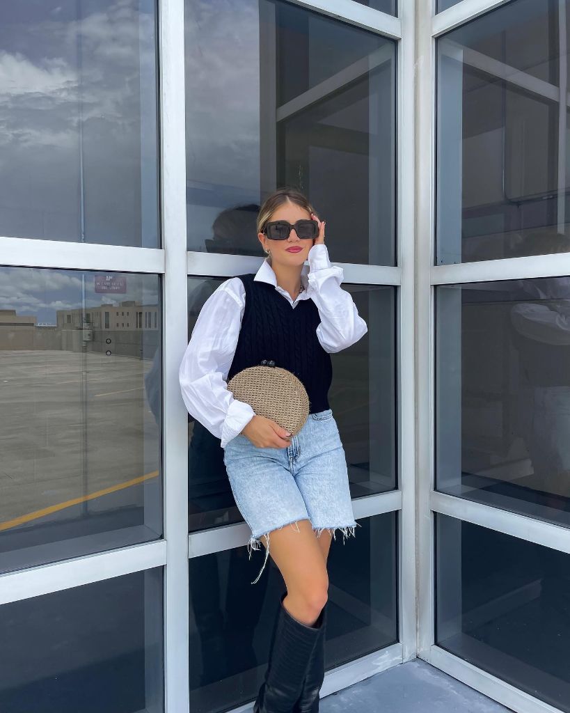 Fall vest outfit with black cable knit sweater vest layered over white button-down shirt, light wash denim bermuda shorts, and tall black boots