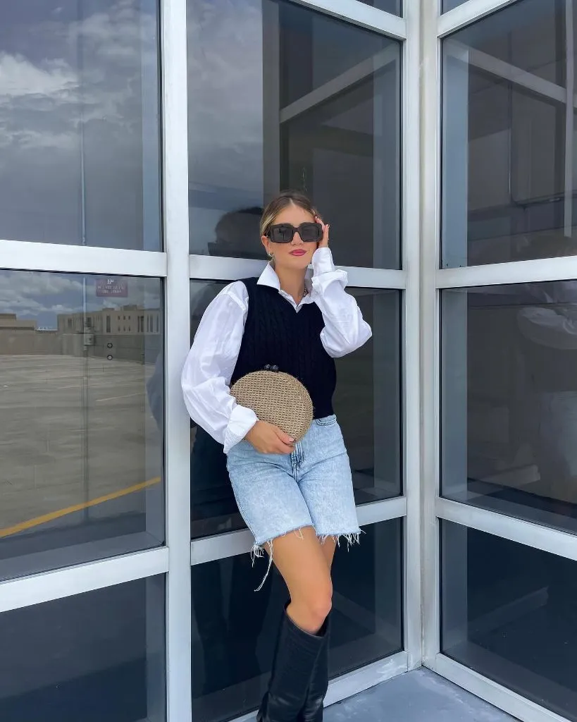 Fall vest outfit with black cable knit sweater vest layered over white button-down shirt, light wash denim bermuda shorts, and tall black boots