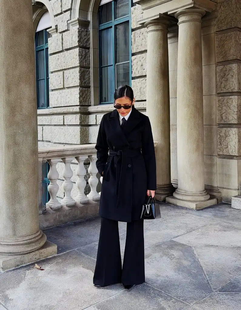 Black belted coat with wide-leg flared trousers and structured mini bag