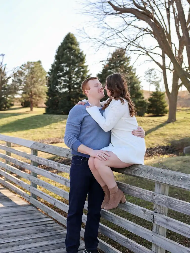 Couple embracing on wooden bridge railing in blue sweater and white dress