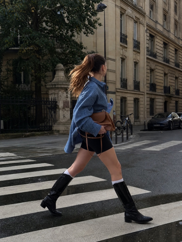 Woman crossing a Paris crosswalk in oversized blue checked shirt, bike shorts, socks and tall black boots