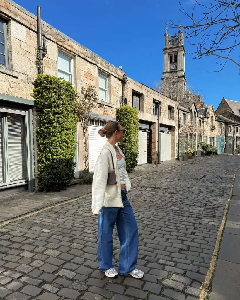 Cream oversized cardigan over white top with wide-leg blue jeans and sneakers on cobblestone Edinburgh street