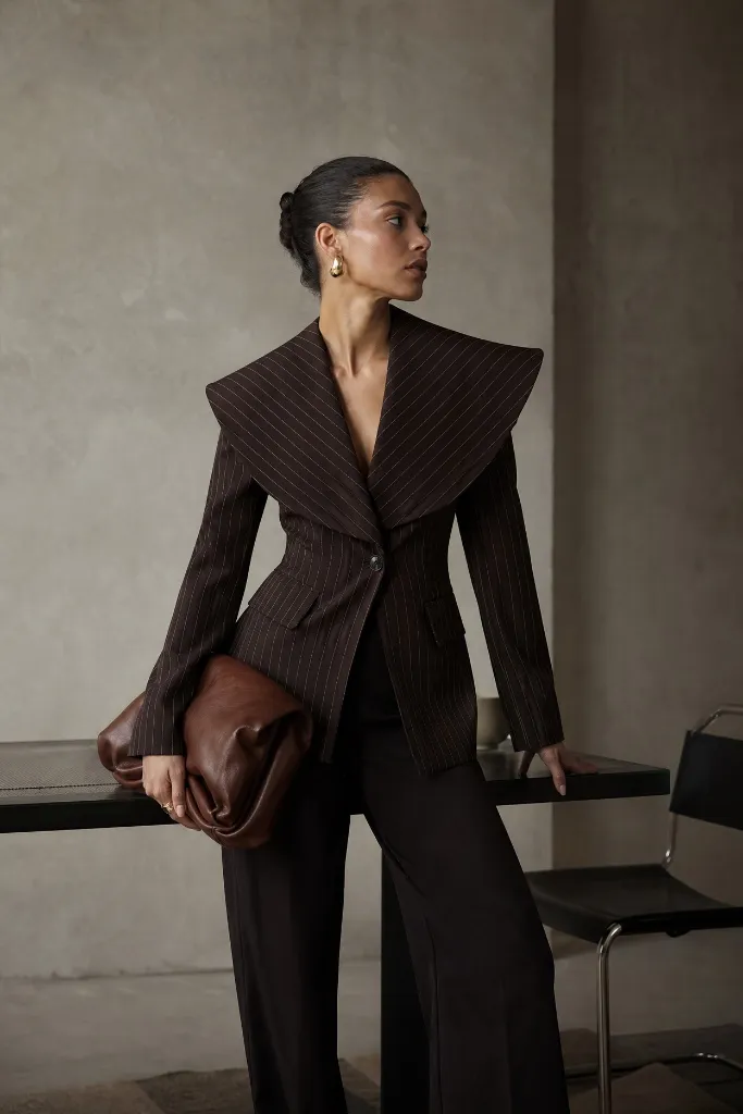 Brown pinstripe blazer with an exaggerated wide portrait collar, nipped waist, black trousers, and a brown leather clutch