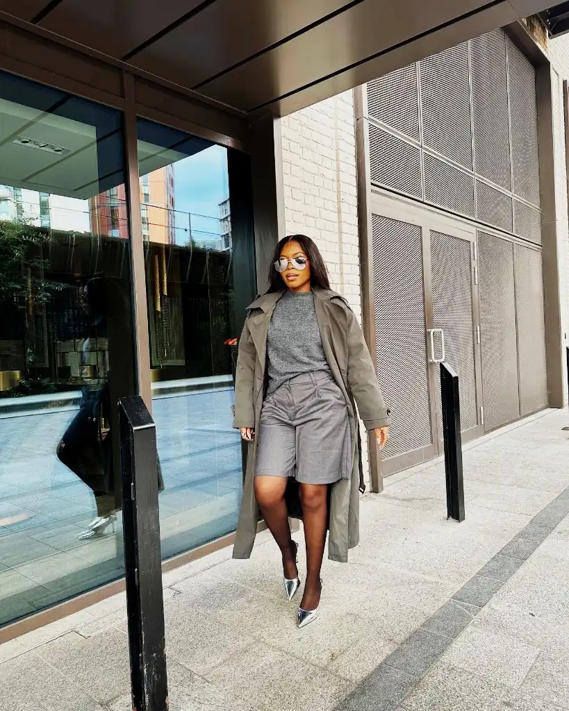 Olive trench coat with grey knit sweater, tailored shorts, and silver metallic mules