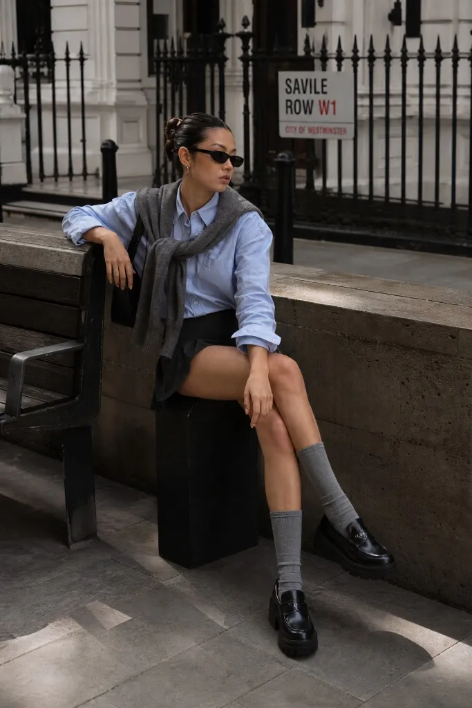 Woman seated in blue button-up shirt, sweater over shoulders, mini skirt, knee socks and loafers