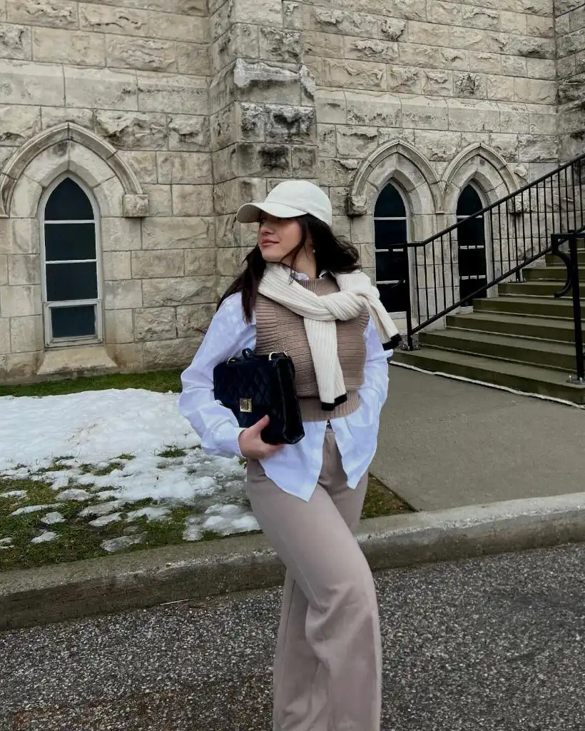 Blush sweater vest over white button-down shirt with taupe trousers, cream sweater draped over shoulders, white cap, and quilted navy bag