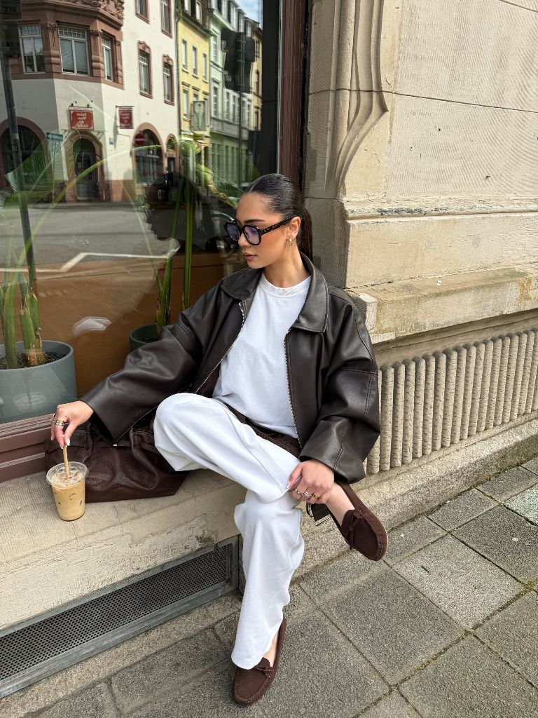 Brown leather oversized jacket with white outfit and moccasin shoes, seated with iced coffee