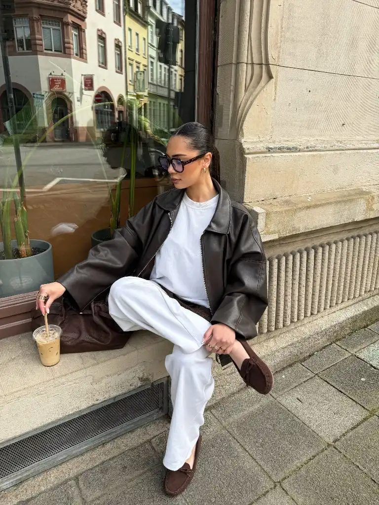 Brown leather oversized jacket with white outfit and moccasin shoes, seated with iced coffee