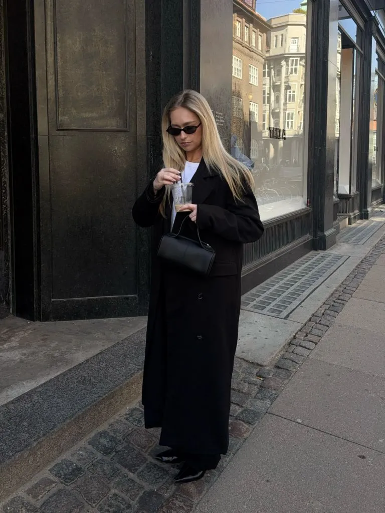 Long black overcoat with white tee, black trousers, black boots and structured bag