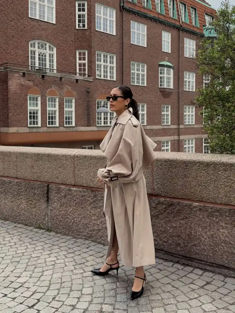 Full-length beige belted trench coat worn as a dress with black kitten heel Mary Janes and gold earrings in Copenhagen