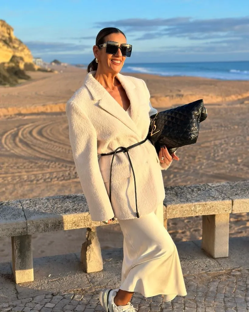 Ivory belted coat with white satin skirt and sneakers on the beach