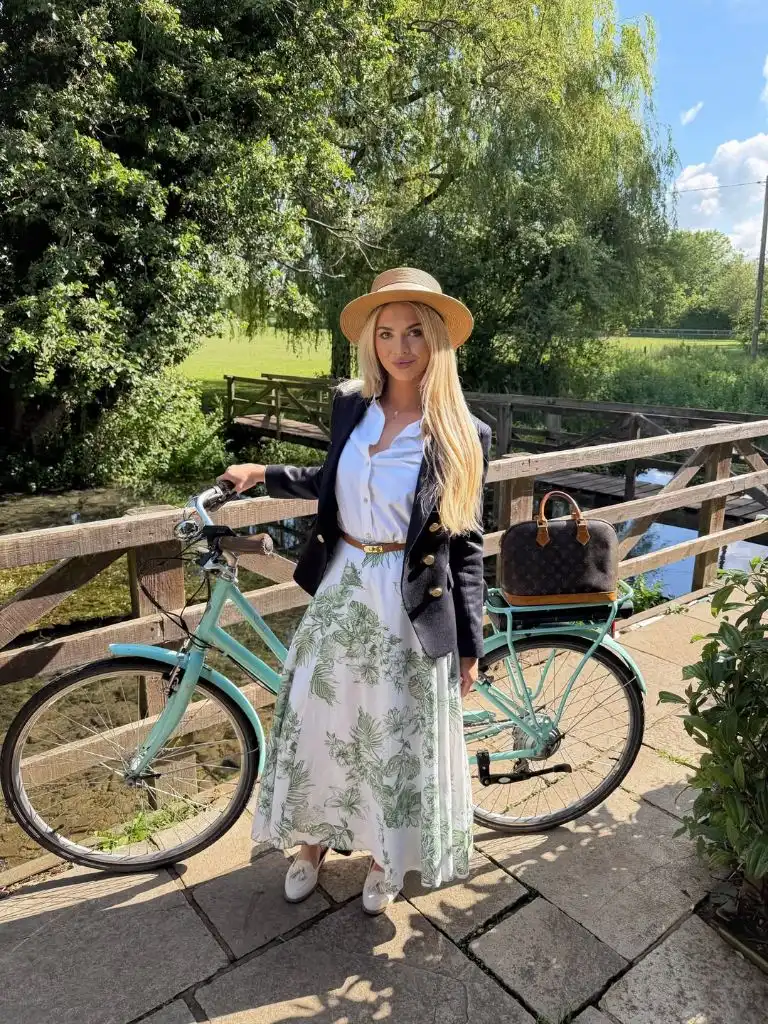 Navy blazer over white button-down shirt with green floral midi skirt, straw hat, and Louis Vuitton bag beside vintage bicycle in English countryside