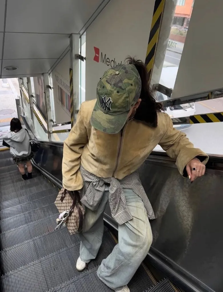 Fall travel outfit with suede bomber jacket, wide-leg jeans, camo cap, and Gucci bag on escalator