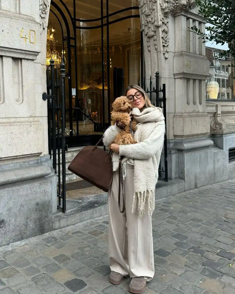 Cream oversized scarf and sweater with beige wide-leg pants and brown tote, holding a puppy