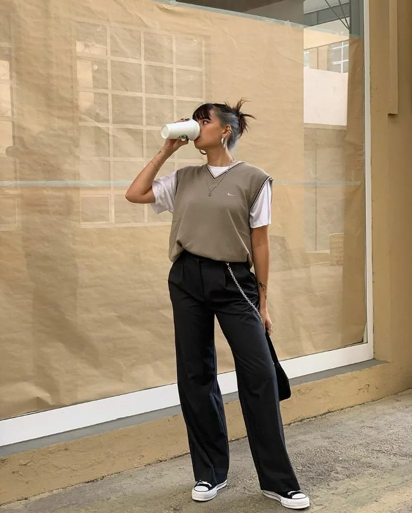 Taupe Nike V-neck sweater vest over white tee with black wide-leg trousers, Converse sneakers, chain crossbody bag, and layered necklaces