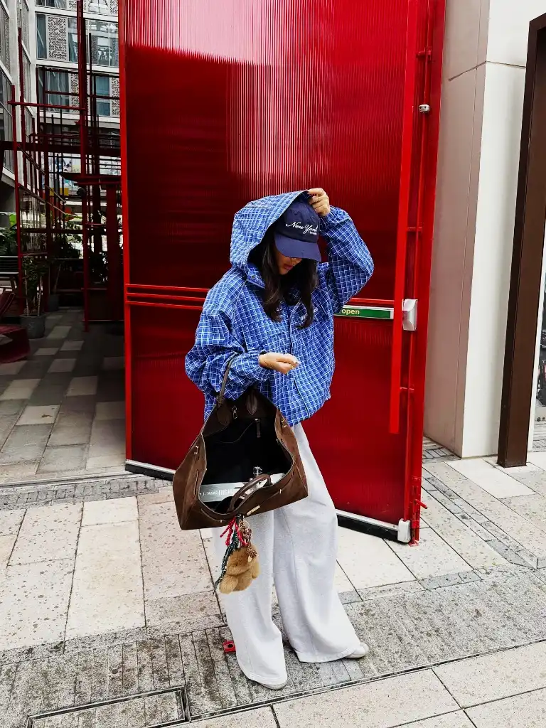 Blue check anorak with grey sweatpants, baseball cap, and brown suede tote