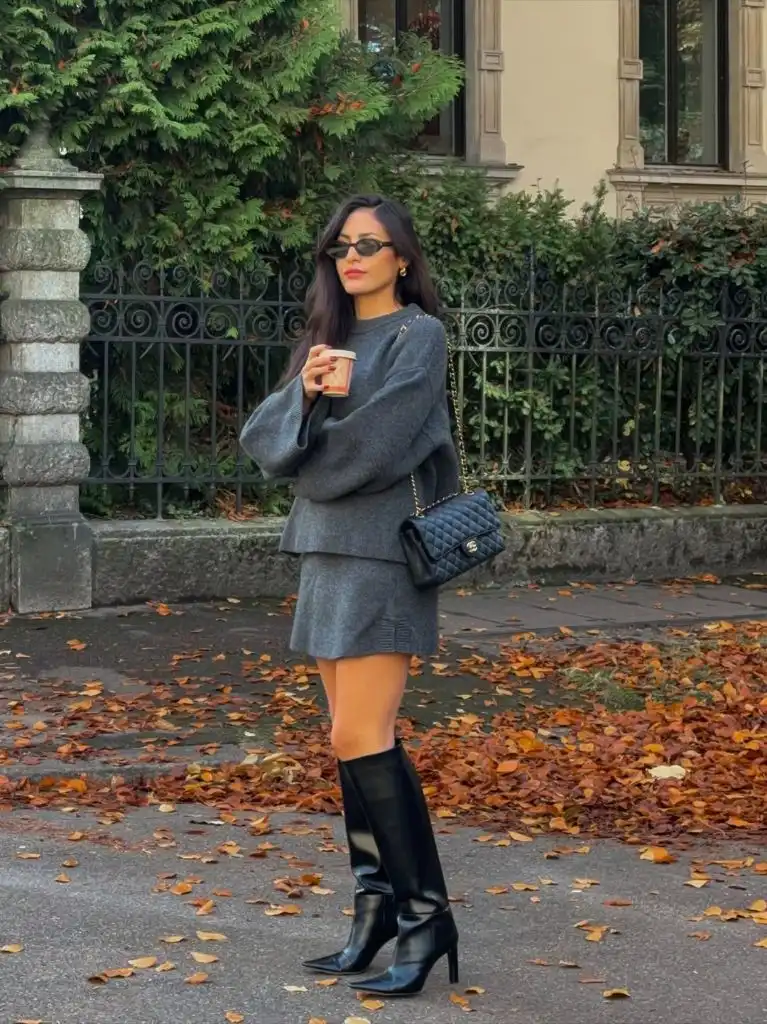 Grey knit sweater and matching mini skirt set with black knee-high heeled boots and Chanel bag among fall leaves