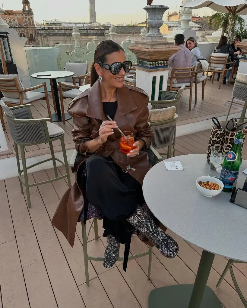 Brown leather trench coat with black trousers and silver boots at a rooftop bar