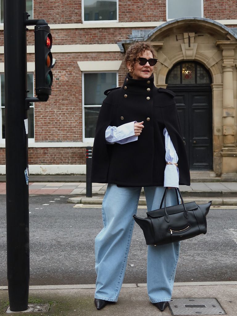 Black cape jacket over blue shirt with wide-leg light denim and black bag