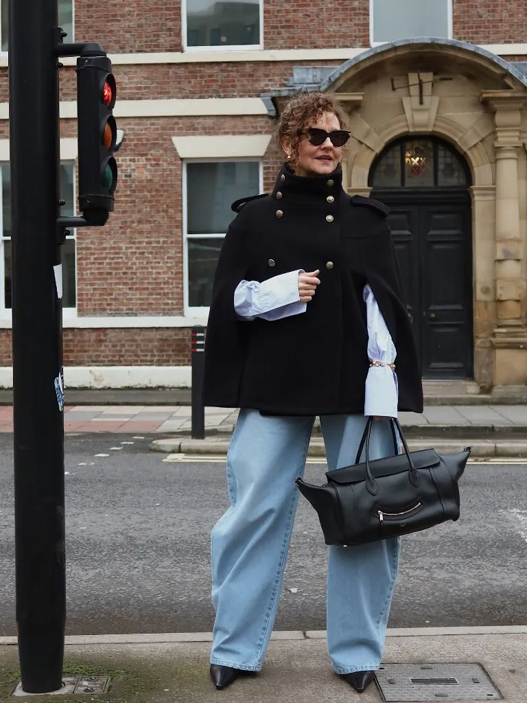Black cape jacket over blue shirt with wide-leg light denim and black bag