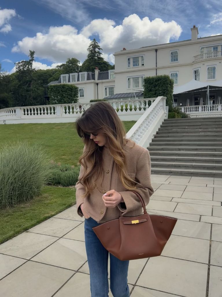 Camel tweed blazer with gold buttons over brown top, blue jeans, and cognac leather tote at grand estate