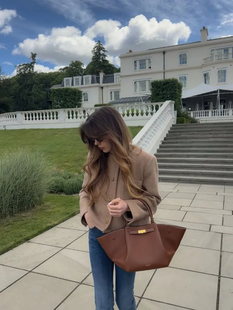 Camel tweed blazer with gold buttons over brown top, blue jeans, and cognac leather tote at grand estate