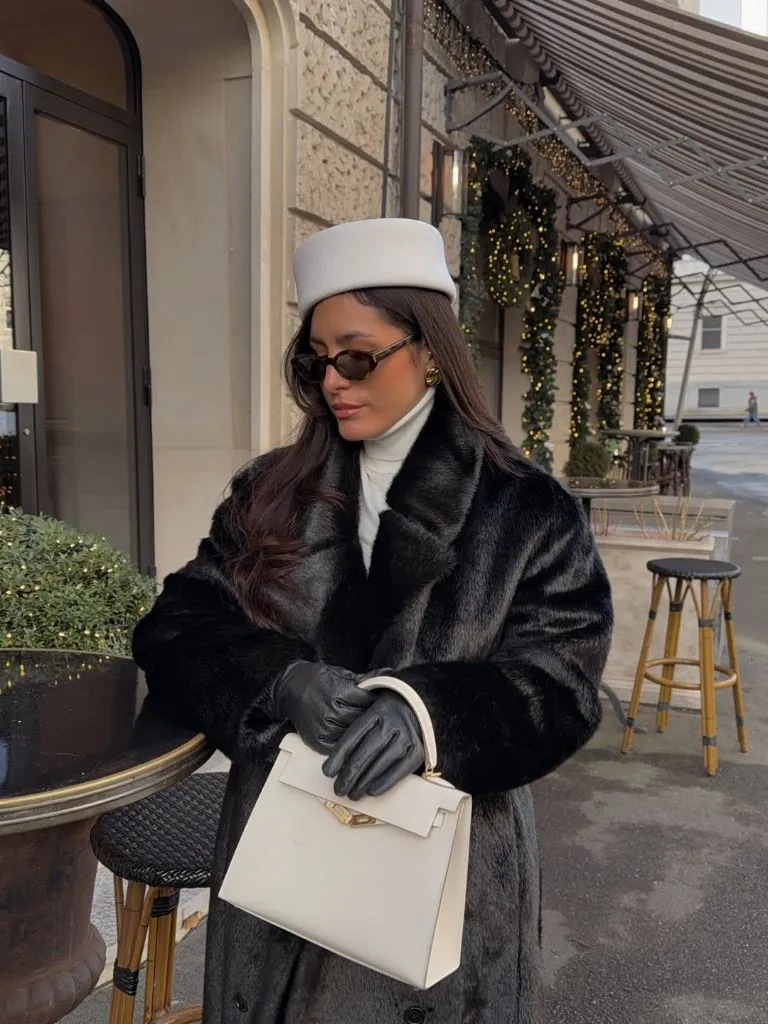 Black faux fur coat with white turtleneck, white pillbox hat, leather gloves, and cream structured handbag at Parisian café
