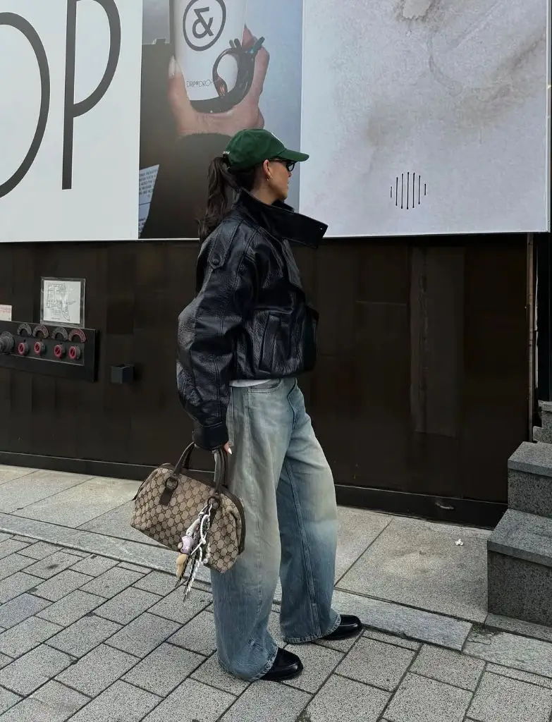 Black leather jacket with wide-leg light wash jeans, green cap, Gucci bag, and black loafers