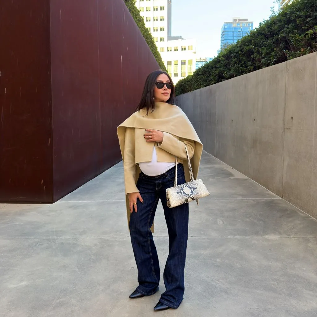 Stylish pregnant woman in camel cape, white top, dark flare jeans, pointed boots, and snakeskin bag