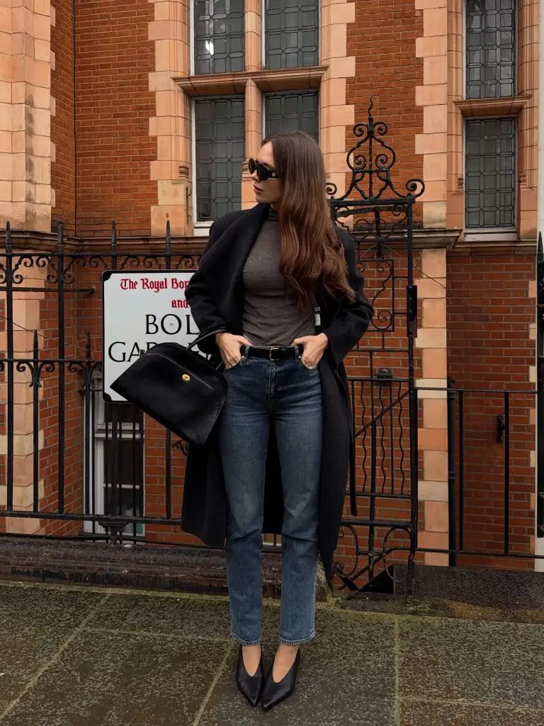 Dark wool coat with brown turtleneck, straight-leg jeans, and pointed black pumps outside a London building