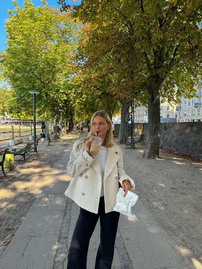 Cream boucle peacoat with black pants and iced coffee on a tree-lined path