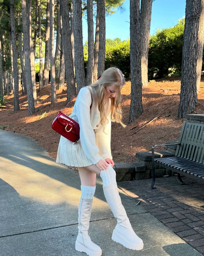 All-white outfit with cream sweater, pleated mini skirt, white platform boots, cable-knit leg warmers, and red patent bag