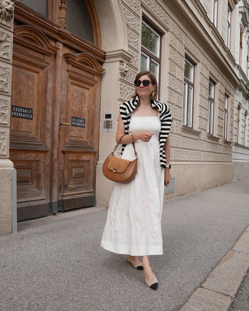White midi dress with black-and-white striped sweater knotted over shoulders, tan saddle bag, and cap-toe flats