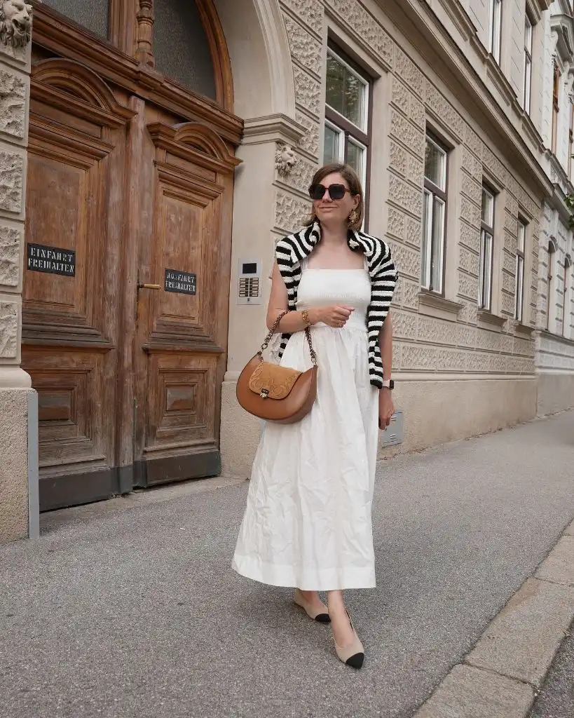 White midi dress with black-and-white striped sweater knotted over shoulders, tan saddle bag, and cap-toe flats