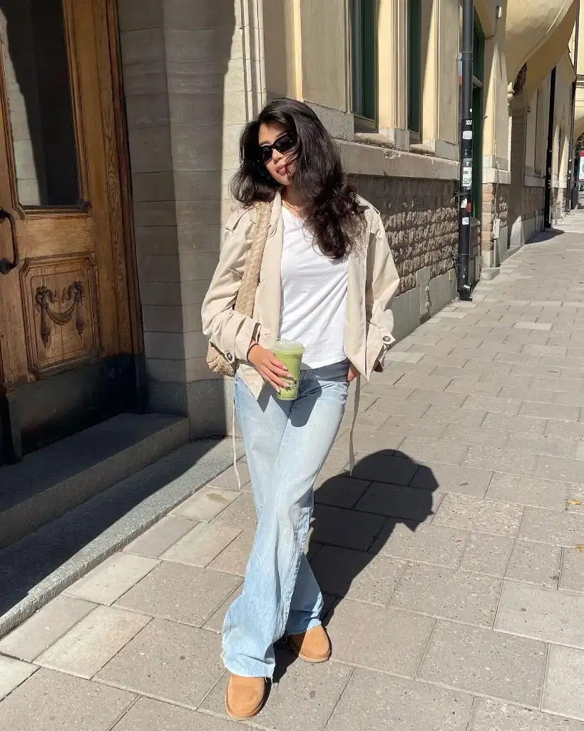 Beige lightweight jacket with white tee, light-wash flared jeans, tan UGG slip-ons, and woven bag