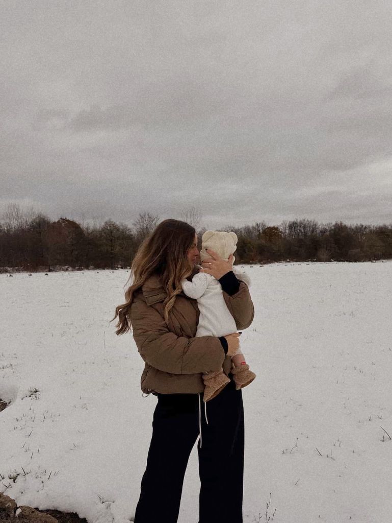 Mom holding baby in snowy field wearing brown puffer