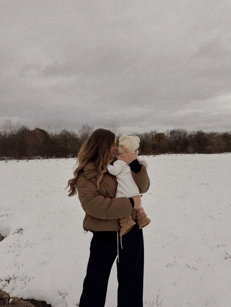Mom holding baby in snowy field wearing brown puffer
