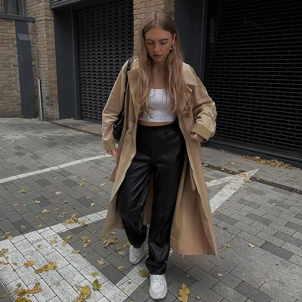 Tan trench coat over white crop top with black faux leather wide-leg trousers and white sneakers on autumn street