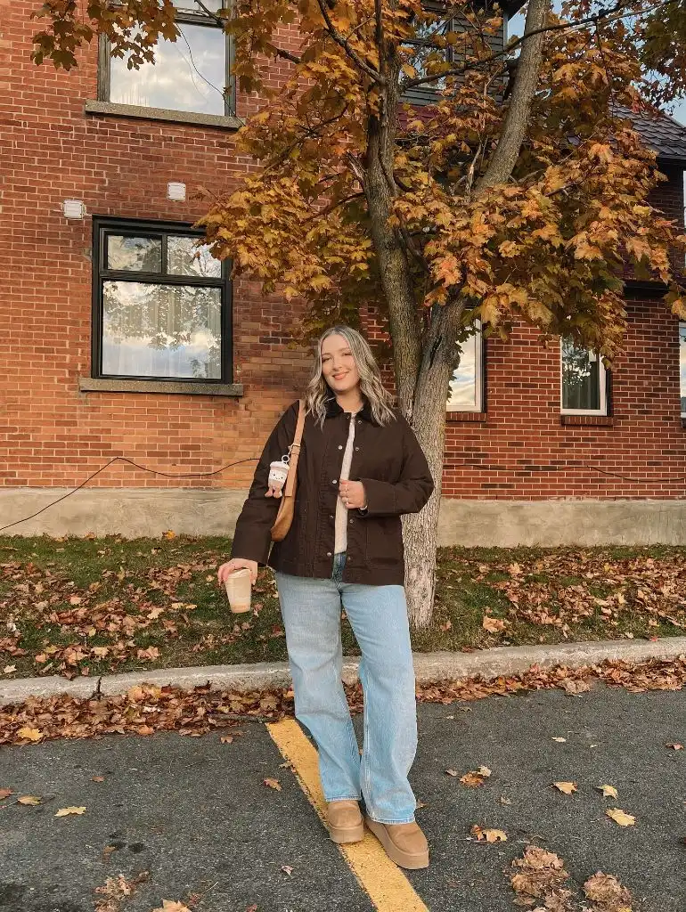 Pregnant woman standing on fall sidewalk in brown chore jacket, wide-leg jeans, and UGG platforms holding iced coffee