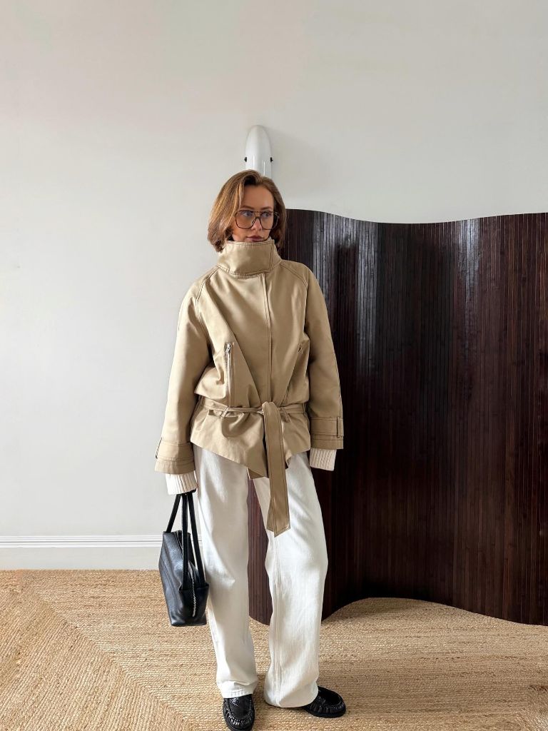 Beige high-collar belted peacoat with white wide-leg trousers and glasses