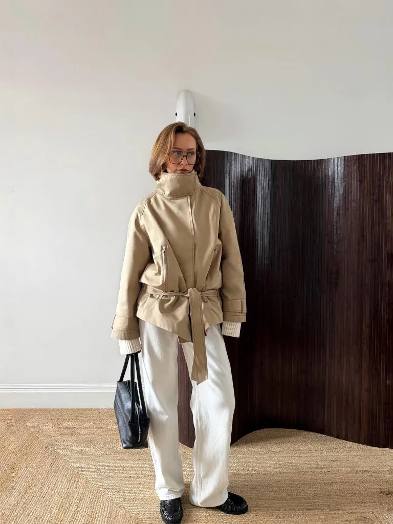 Beige high-collar belted peacoat with white wide-leg trousers and glasses