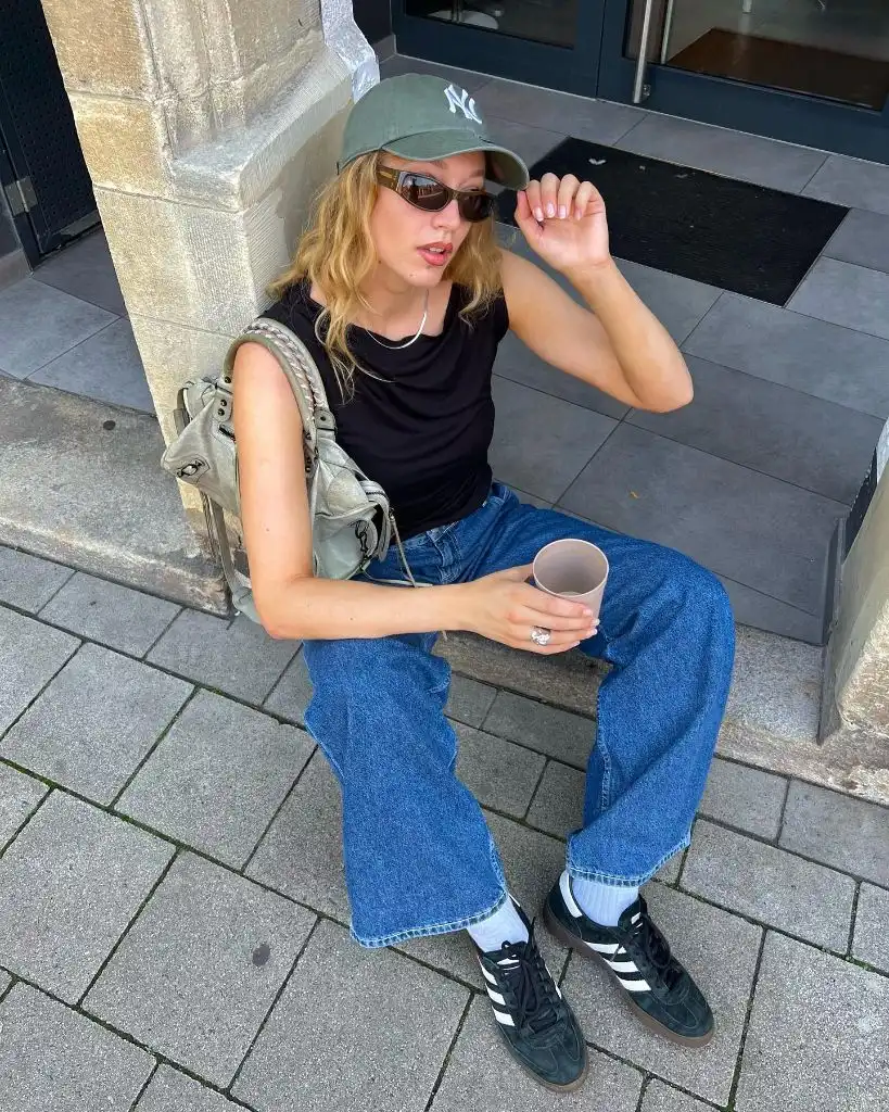 Black tank top with blue jeans, NYC cap, Adidas Samba sneakers, and sage green backpack