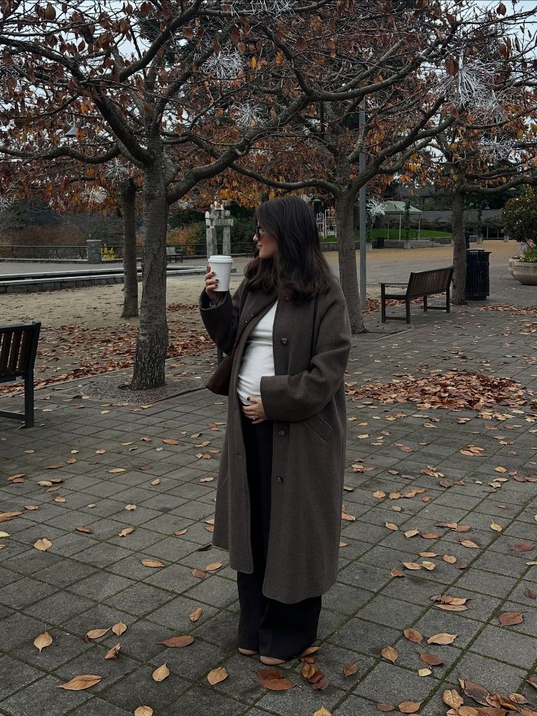 Pregnant woman in brown wool overcoat, white tee, black pants, holding coffee in autumn park