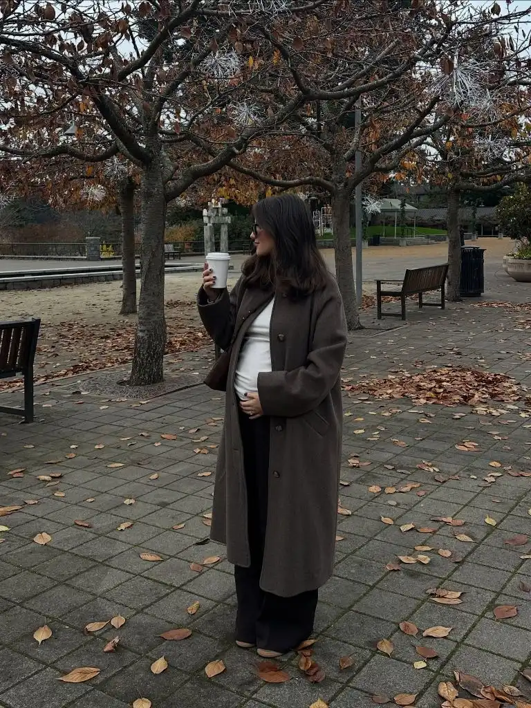 Pregnant woman in brown wool overcoat, white tee, black pants, holding coffee in autumn park