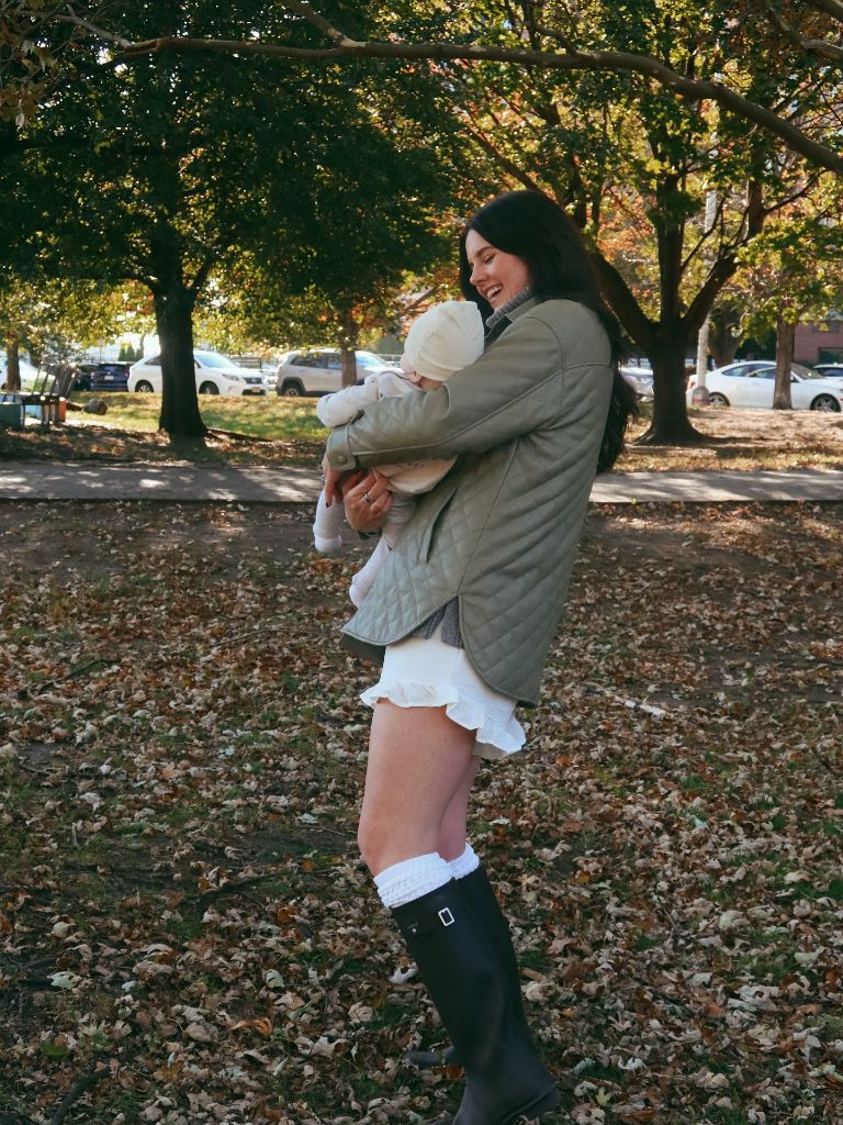 Mom holding baby in park wearing quilted jacket and tall boots